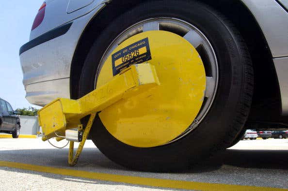 A yellow, boot-style trailer wheel lock with “City of Chicago” written on the side sits on the right, rear wheel of a parked silver sedan. 右边是一辆停放着的银色轿车的后轮,上面有一个黄色的后轮锁,侧面写着“芝加哥市”。
