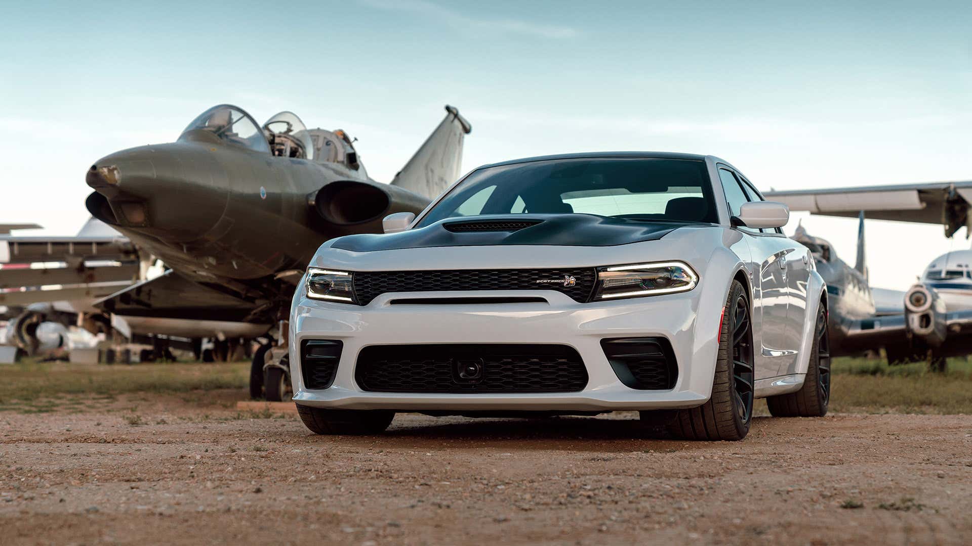A white 2021 Dodge Charger Widebody Scat Pack rests next to a jet fighter. 白色2021年道奇充电器宽体猫包休息旁边的喷气战斗机。