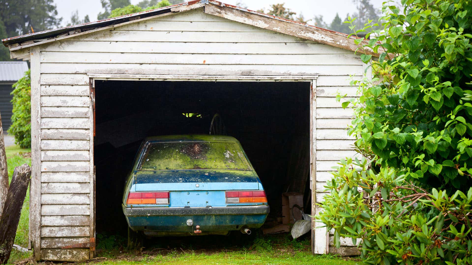An abandoned European car is seen in a small garage with worn white paint. 一辆废弃的欧洲汽车停在一个小车库里,白色油漆已经磨损。